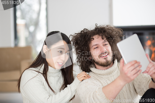 Image of multiethnic couple using tablet computer in front of fireplace