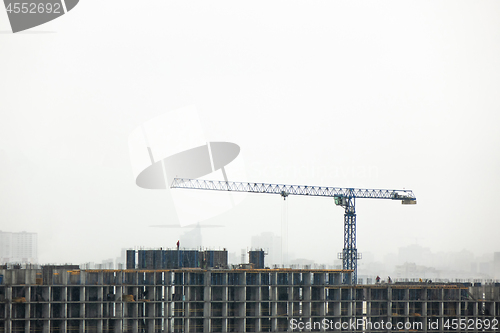 Image of Construction crane against the cloudy background and the silhouettes of buildings in the misty sky.