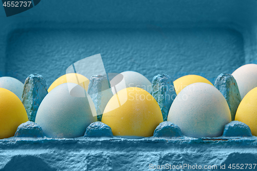 Image of Close-up multicolored painted in yellow blue eggs in a blue cardboard box around blue background with copy space. Easter composition.