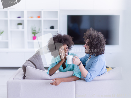 Image of multiethnic couple sitting on sofa at home drinking coffe