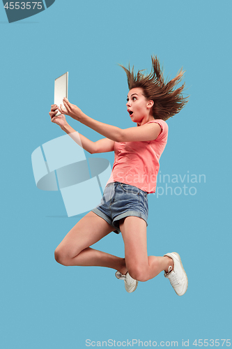 Image of Image of young woman over blue background using laptop computer or tablet gadget while jumping.