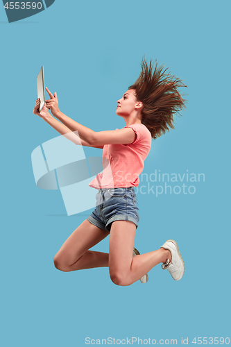 Image of Image of young woman over blue background using laptop computer or tablet gadget while jumping.