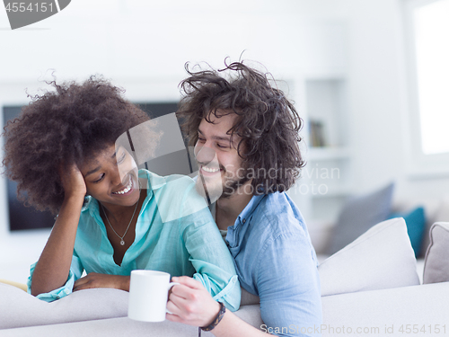 Image of multiethnic couple sitting on sofa at home drinking coffe