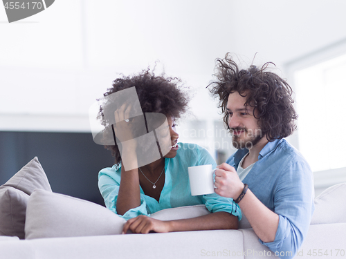 Image of multiethnic couple sitting on sofa at home drinking coffe