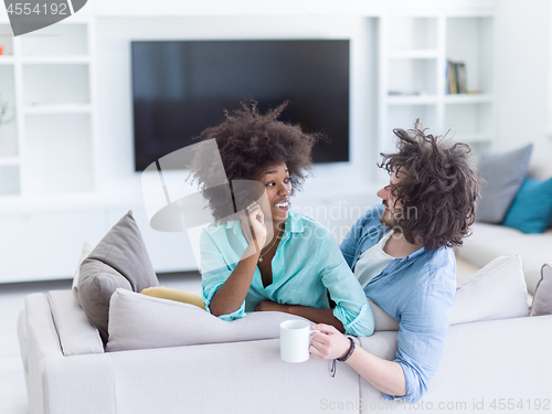 Image of multiethnic couple sitting on sofa at home drinking coffe