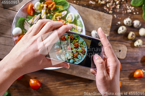 Image of Hands of a girl with a phone make a photo of a freshly prepared salad. Food blogging concept. Top view