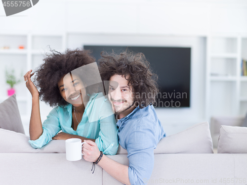 Image of multiethnic couple sitting on sofa at home drinking coffe