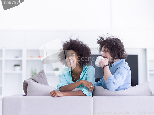 Image of multiethnic couple sitting on sofa at home drinking coffe