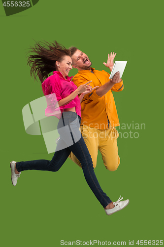 Image of Jump of young couple over green studio background using laptop or tablet gadget while jumping.