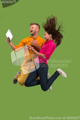 Image of Jump of young couple over green studio background using laptop or tablet gadget while jumping.