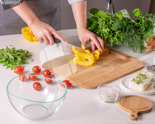 Image of A woman\'s hand slices a yellow pepper on a wooden board on a white kitchen table. Preparation of salad