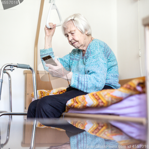 Image of Elderly 96 years old woman reading phone message while sitting on medical bed supporting her by holder.