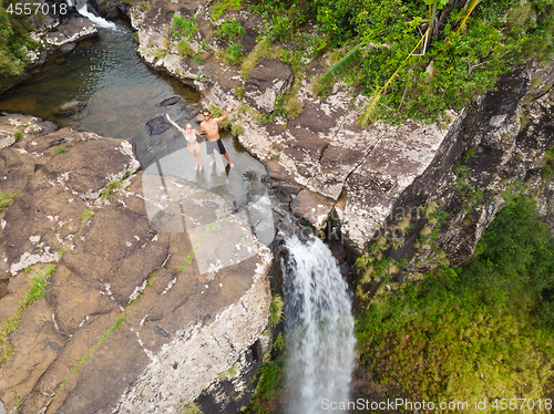 Image of Aerial top view of travel couple waving to drone, standing on the edge of 500 feet waterfall in the tropical island jungle of Black river gorges national park on Mauritius island