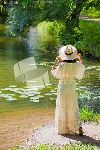 Image of girl in a white dress and hat on the shore of a pond with water-