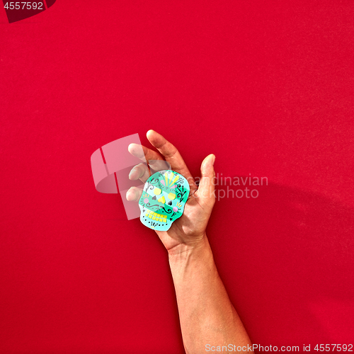 Image of The hand of a man holds a handcraf paper skull Calaveras attributes of the Mexican holiday Calaca on a red background with space for text and reflection of shadows. Halloween. Flat lay