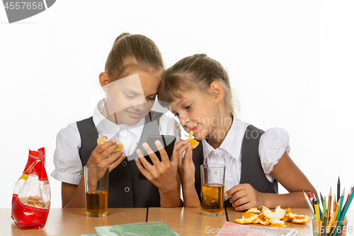 Image of Two schoolgirls at a break look at the screen of a smartphone, and eat an orange and drink juice
