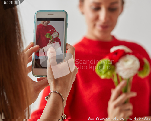 Image of The girl takes a photo on the phone of a young pretty woman with a bouquet of fresh flowers on a gray background.