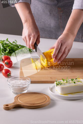 Image of The girl\'s hand cuts pieces of pepper on a wooden board, cheese, and tomatoes on a white kitchen table. Preparation of salad