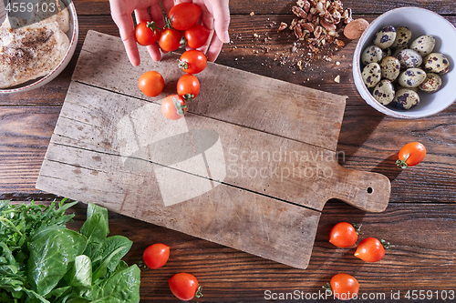 Image of The hands of the woman put ripe tomatoes on a wooden board with quail eggs, pieces of nuts and copy space. The concept of cooking salad. Flat lay