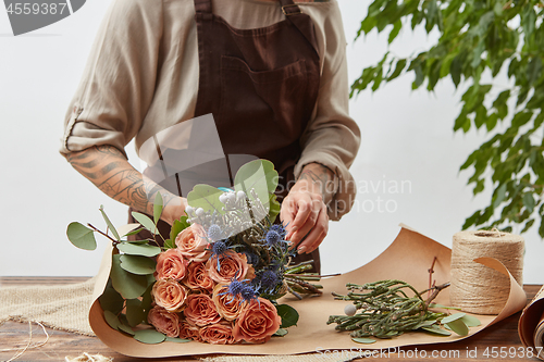 Image of Florist at workspace. Woman\'s hands with tattoo are making bouquet from fresh natural flowers coral living color with decorative leaves on a wooden table step by step.