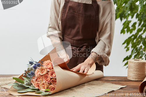 Image of Florist woman is wrapping fresh rose bouquet in a decorative paper on a wooden table on a light wall background. Process step by step. Mother\'s Day holiday.