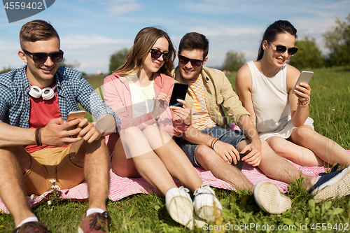 Image of smiling friends with smartphones sitting on grass