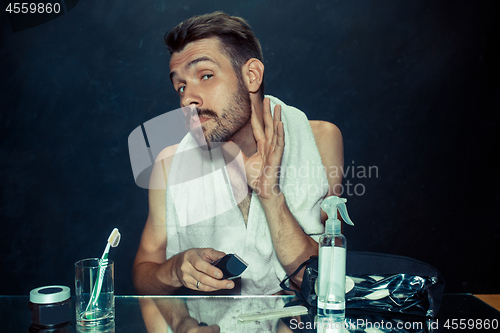 Image of young man in bedroom sitting in front of the mirror scratching his beard