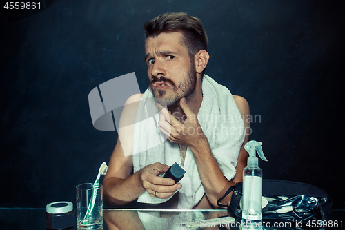 Image of young man in bedroom sitting in front of the mirror scratching his beard