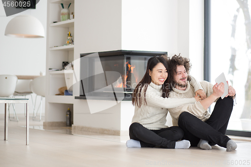 Image of multiethnic couple using tablet computer in front of fireplace