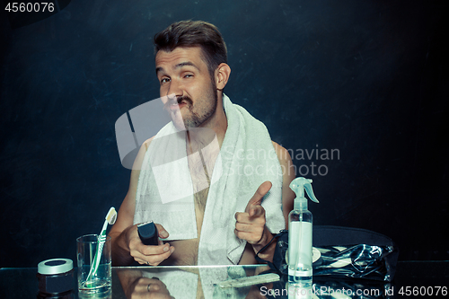 Image of young man in bedroom sitting in front of the mirror scratching his beard