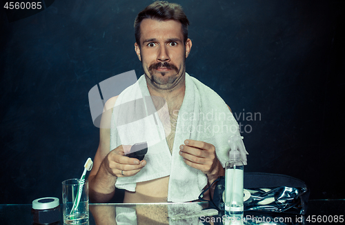 Image of young man in bedroom sitting in front of the mirror scratching his beard