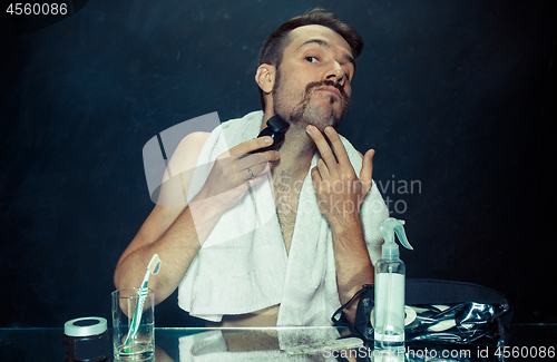 Image of young man in bedroom sitting in front of the mirror scratching his beard