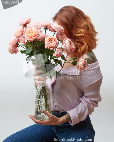 Image of Red-haired girl with a tattoo and a beautiful pink bouquet of roses in a vase on a gray background with copy space. Mothers Day.