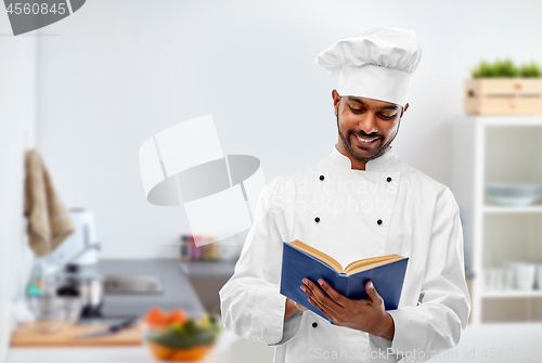 Image of happy male indian chef reading cookbook at kitchen