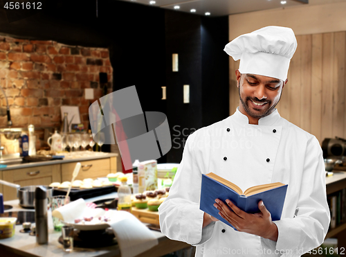 Image of happy male indian chef reading cookbook at kitchen