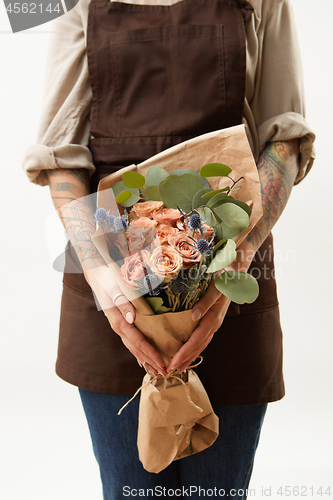 Image of Woman in a brown apron is holding summer wonderful fresh roses, eryngium and green leaves as a greeting bunch on a light background. Greeting card. Copy space.