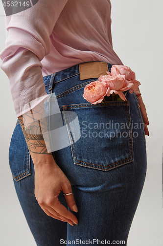 Image of Young pretty woman dressed in blue jeans and pink jacket with fresh roses in a back pocket and her hand tattoo on a gray background, copy space.