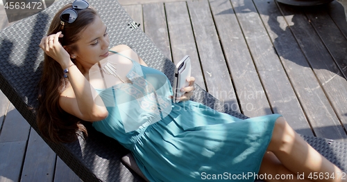 Image of Woman in blue dress resting in lounge chair and using smartphone