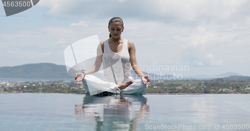 Image of Serene woman practicing yoga in lotus posture poolside against sky