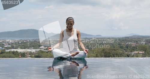 Image of Calm woman practicing yoga in lotus posture poolside against resort town