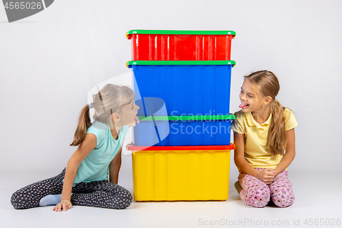 Image of Two girls peek from different sides of large plastic boxes and show each other their tongues