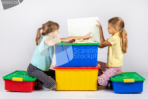 Image of Two girls sit on large toy boxes and look at the toy assembly instructions