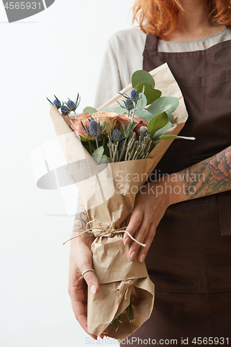 Image of Female in a brown apron with red hair hold nice bouquet from living coral colored roses and eryngium on a light