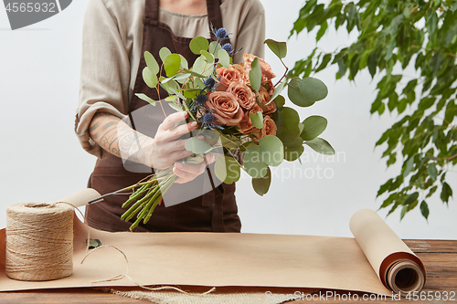 Image of Young woman florist is making bouquet with fresh flowers roses living coral color at the table with paper and rope. Process step by step. Concept floral shop.
