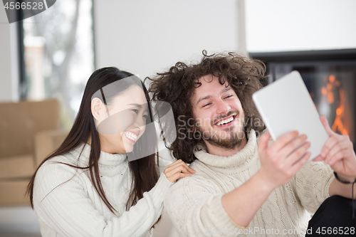 Image of multiethnic couple using tablet computer in front of fireplace