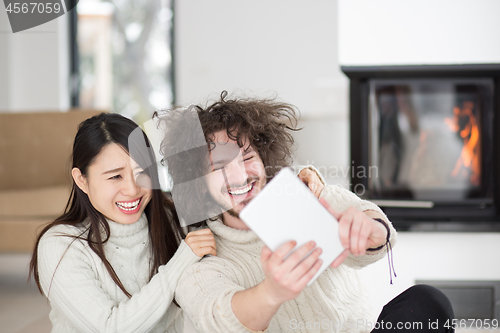 Image of multiethnic couple using tablet computer in front of fireplace