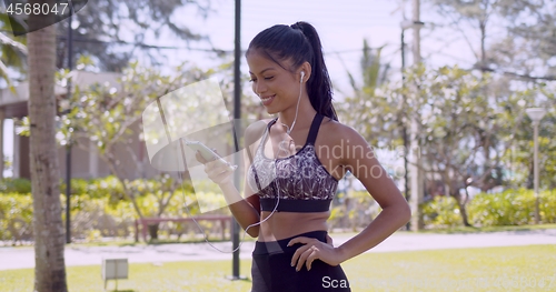 Image of Cheerful Asian woman in earphones browsing smartphone in hand in park