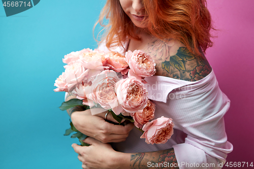 Image of Attractive red-haired girl with tattoo on shoulders and hands holds a bunch of coral roses on a blue-pink background. A gift for Mother\'s Day.