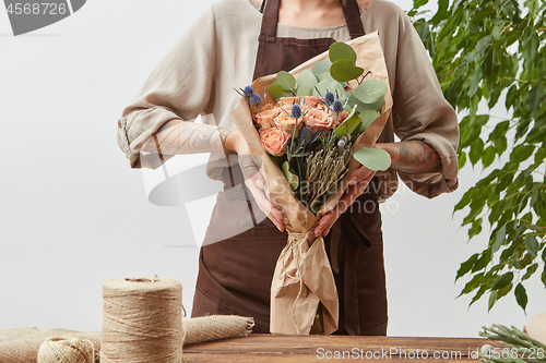 Image of Girl\'s florist hands hold fresh natural bouquet from fragrant flowers living coral color roses, green leaf in a paper on a background of gray wall, copy space.