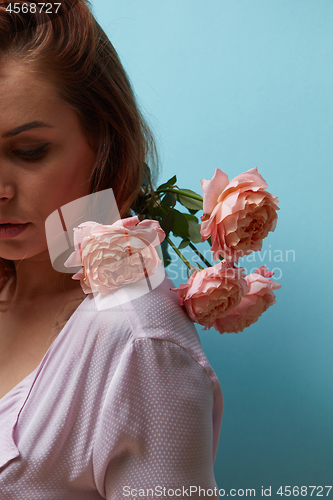 Image of Red-haired girl with fresh pink rose decorating shoulders around the blue background with copy space. Mother\'s day concept